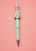 Handmade mint green beaded ballpoint pen with mauve pink silicone bead, white gold iridescent floral silicone bead, mint green beach gnome resin charm with polka dot hat black bow tie and pink seashell on mint green pen body with silver tip on pink background