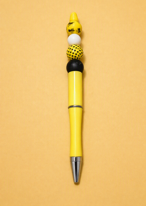 Handmade bright yellow beaded ballpoint pen with matte black silicone bead, yellow black polka dot silicone bead, white silicone bead, yellow honeybee print silicone bead on bright yellow pen body with silver accent on golden yellow background